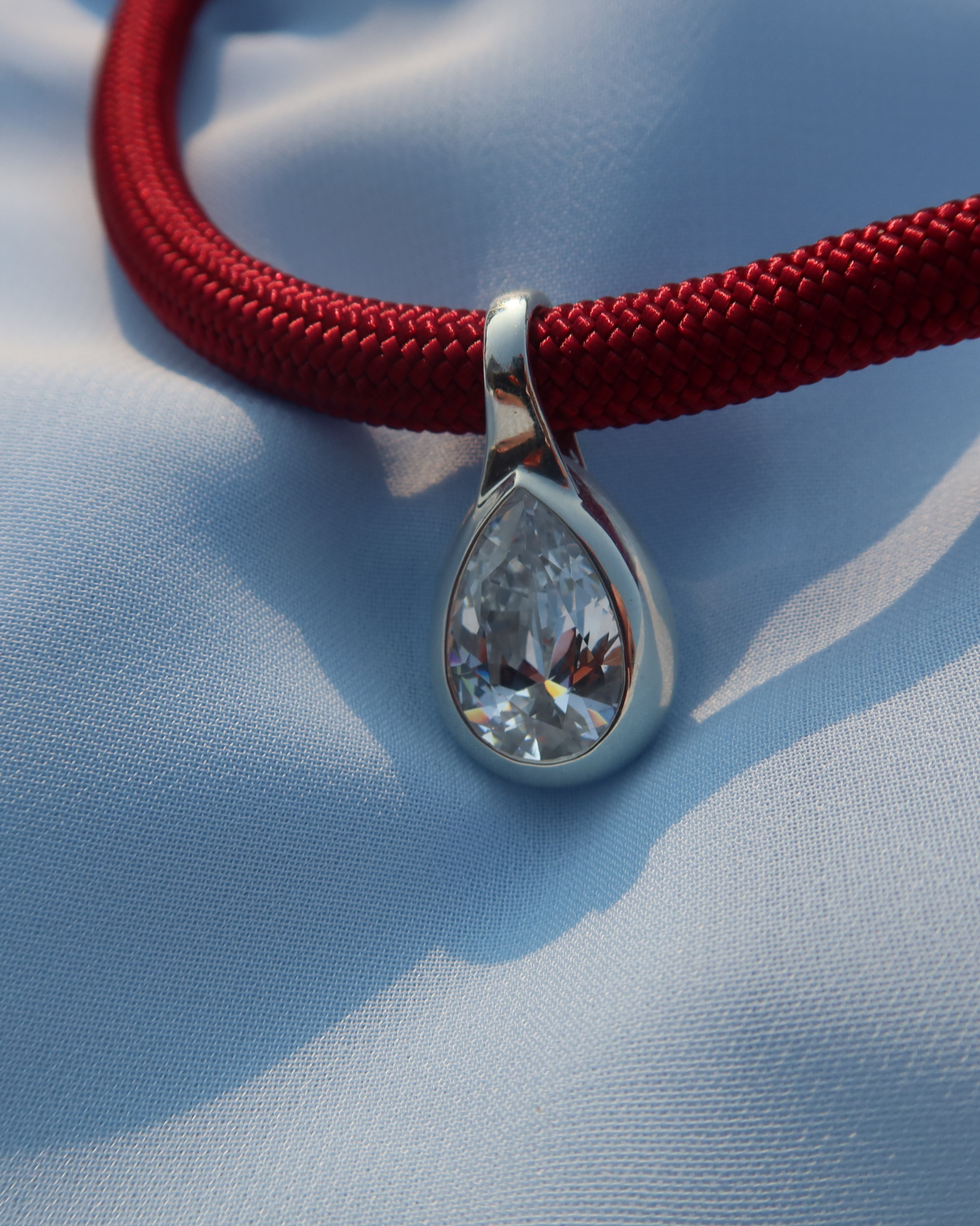 Teardrop-shaped silver pendant with a clear gemstone on a red cord against a blue fabric background
