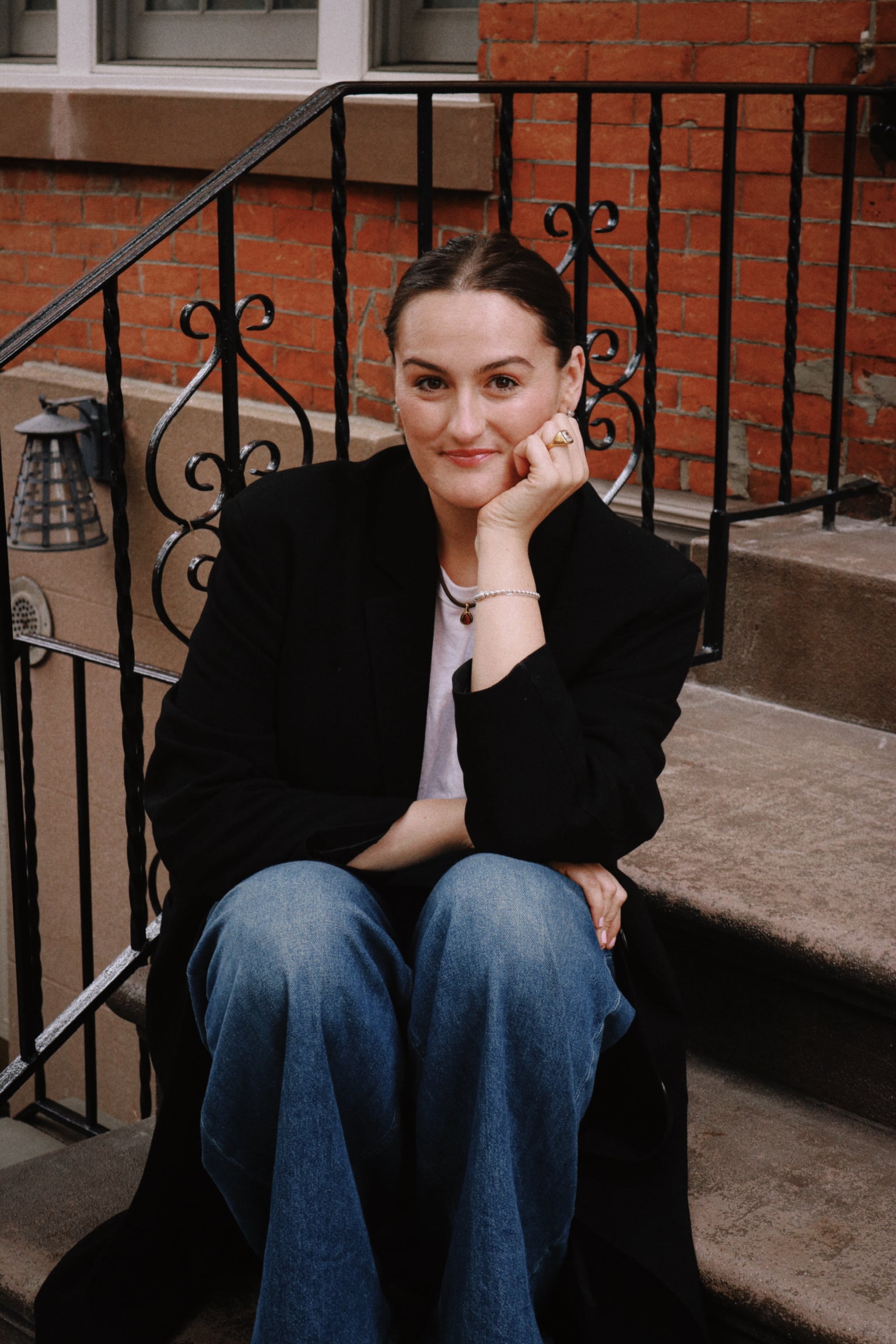 Laura Sneed sitting on steps with a black jacket and blue jeans, brick building in the background